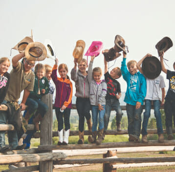 Group of kids sitting on a fence all holding their cowboy hats in the air